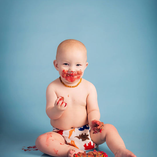 A young baby with a cake on a blue background in a diaper with colorful dinosaurs and cake all over his face.  This image demonstrates the Pure Retouch Photoshop Technique.  There is no line or blemish on the seamless background paper and the skin tone of the subject is very even and smooth and there are no under eye circles or blemishes, but maintains a realistic look.