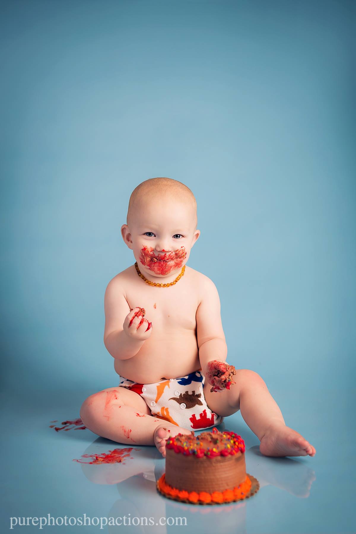 A young baby with a cake on a blue background in a diaper with colorful dinosaurs and cake all over his face.  This image demonstrates the Pure Retouch Photoshop Technique.  There is no line or blemish on the seamless background paper and the skin tone of the subject is very even and smooth and there are no under eye circles or blemishes, but maintains a realistic look.